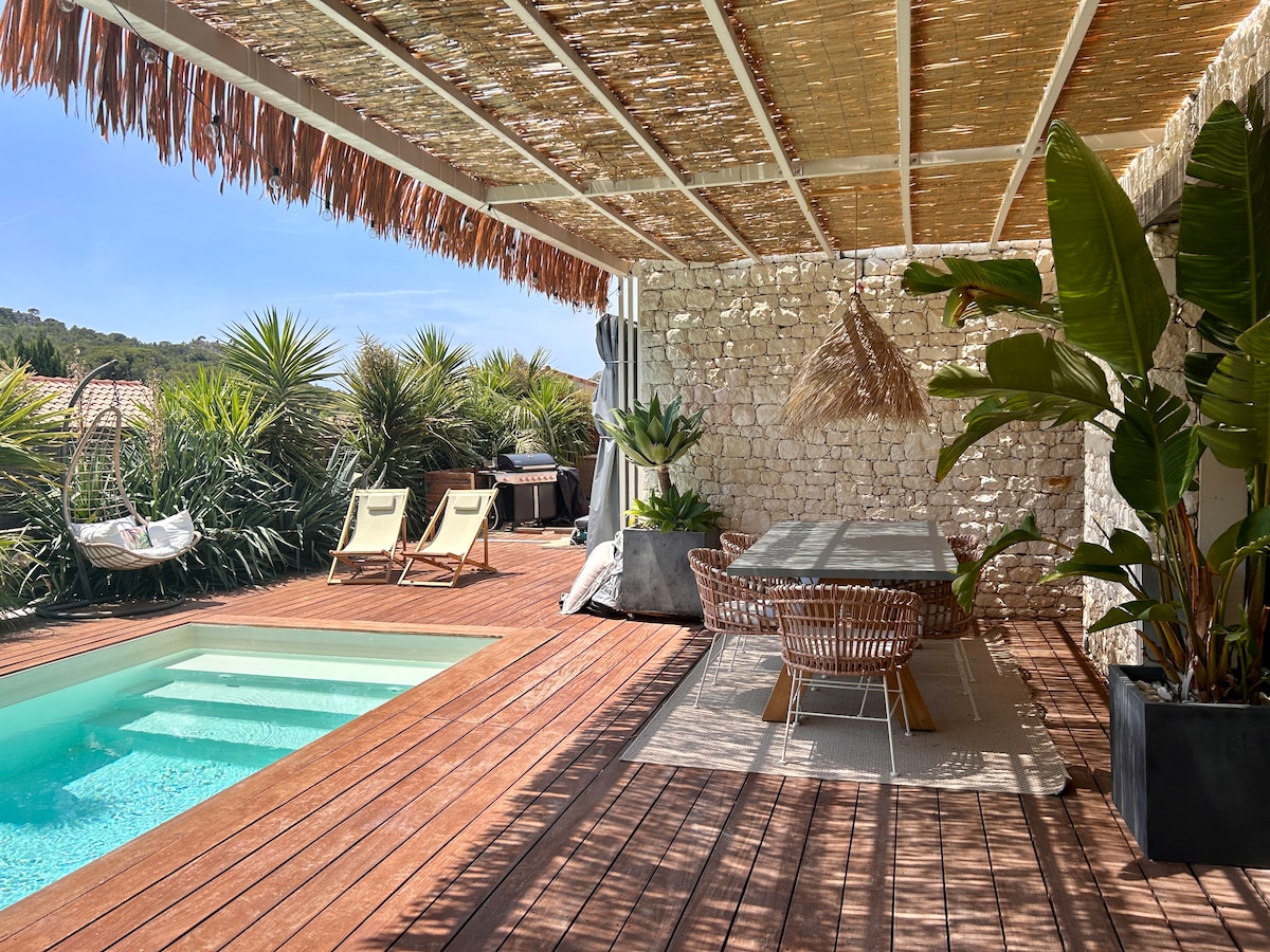An outdoor terrace features a refreshing pool with steps leading down to the water. A dining area with a large table and wicker chairs is shaded by a roof of natural materials. Two lounge chairs offer a space for relaxation, surrounded by lush greenery.