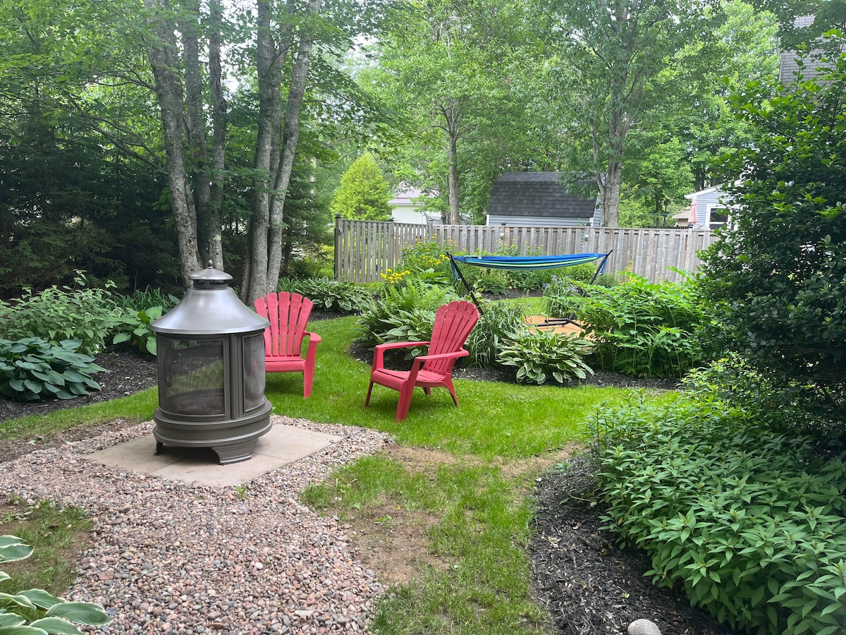 A private outdoor space is featured, with a circular gravel area surrounding a metal fire pit. Two red Adirondack chairs are positioned nearby, complemented by lush greenery and a hammock between the trees. A wooden table is visible in the background, enhancing the garden's charm.