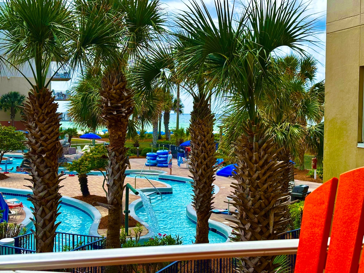 A view from the balcony showcases a vibrant pool area surrounded by palm trees. The scene features a lazy river, lounge chairs, and water play structures. The Gulf of Mexico can be seen in the background, adding to the relaxing atmosphere.