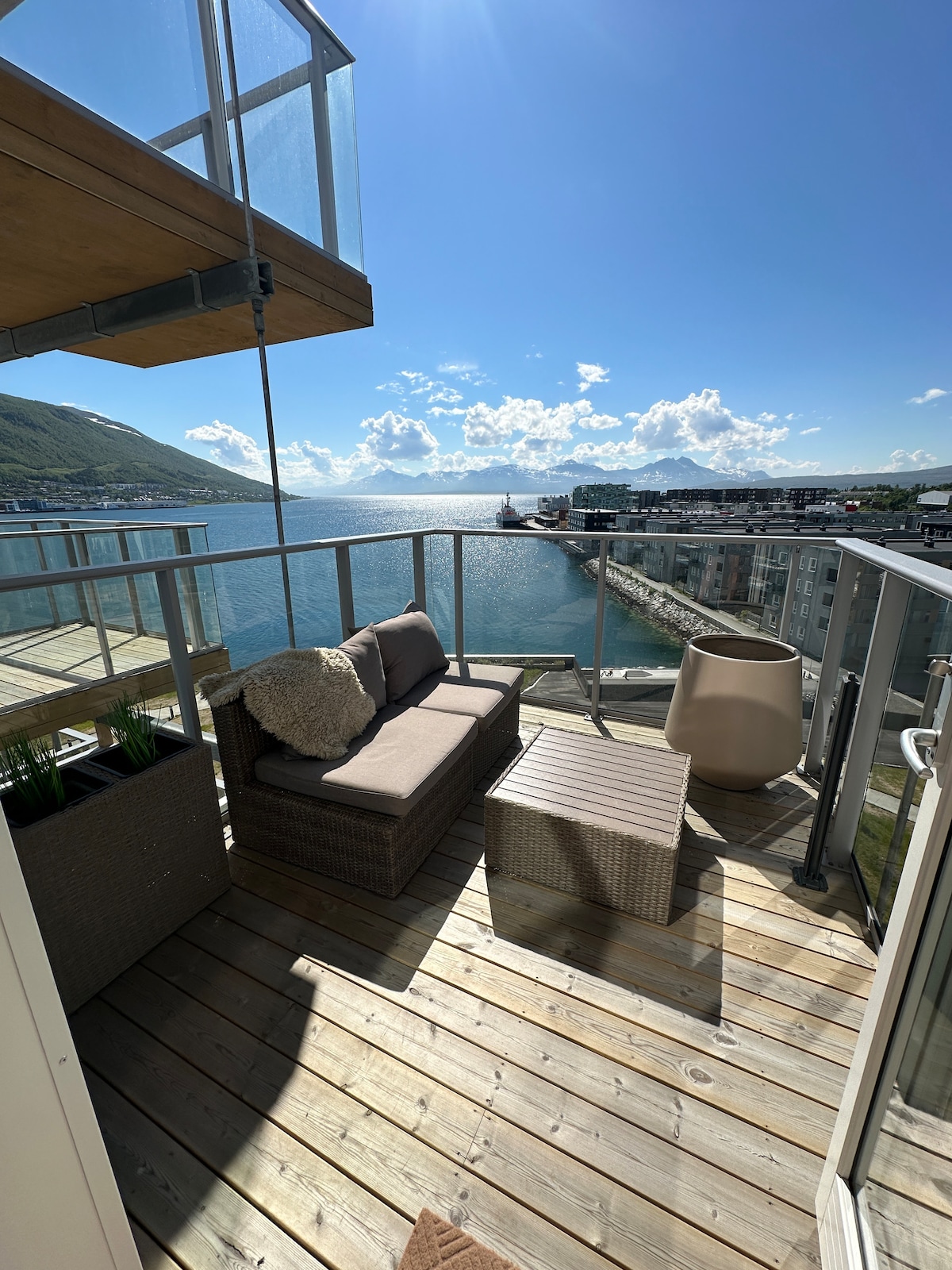 A private balcony offers a cozy seating area with an outdoor sofa and a small coffee table. Natural wood decking complements the surrounding glass railing. The view includes glistening water and distant mountains under a clear blue sky.