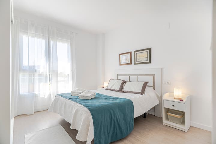 Chumba kikuu cha kulala kilicho na kitanda mara mbili, kitanda cheupe, blanketi la turquoise, taulo zilizokunjwa, kabati la mlango linaloteleza, meza mbili zilizo na taa, michoro kwenye ubao wa kichwa na dirisha kubwa lenye mapazia ya kupendeza ambayo huleta mwanga wa asili kwenye chumba.