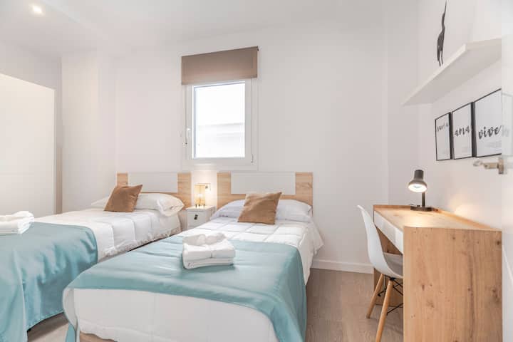 Chumba cha kulala kilicho na vitanda viwili vya mtu mmoja, vitanda vyeupe, mablanketi ya turquoise, mito ya kahawia, taulo zilizokunjwa, kabati la mlango linaloteleza, dawati lenye taa, kiti cheupe, rafu ya mapambo, kioo na dirisha lenye kipofu ambalo hutoa mwanga wa asili.