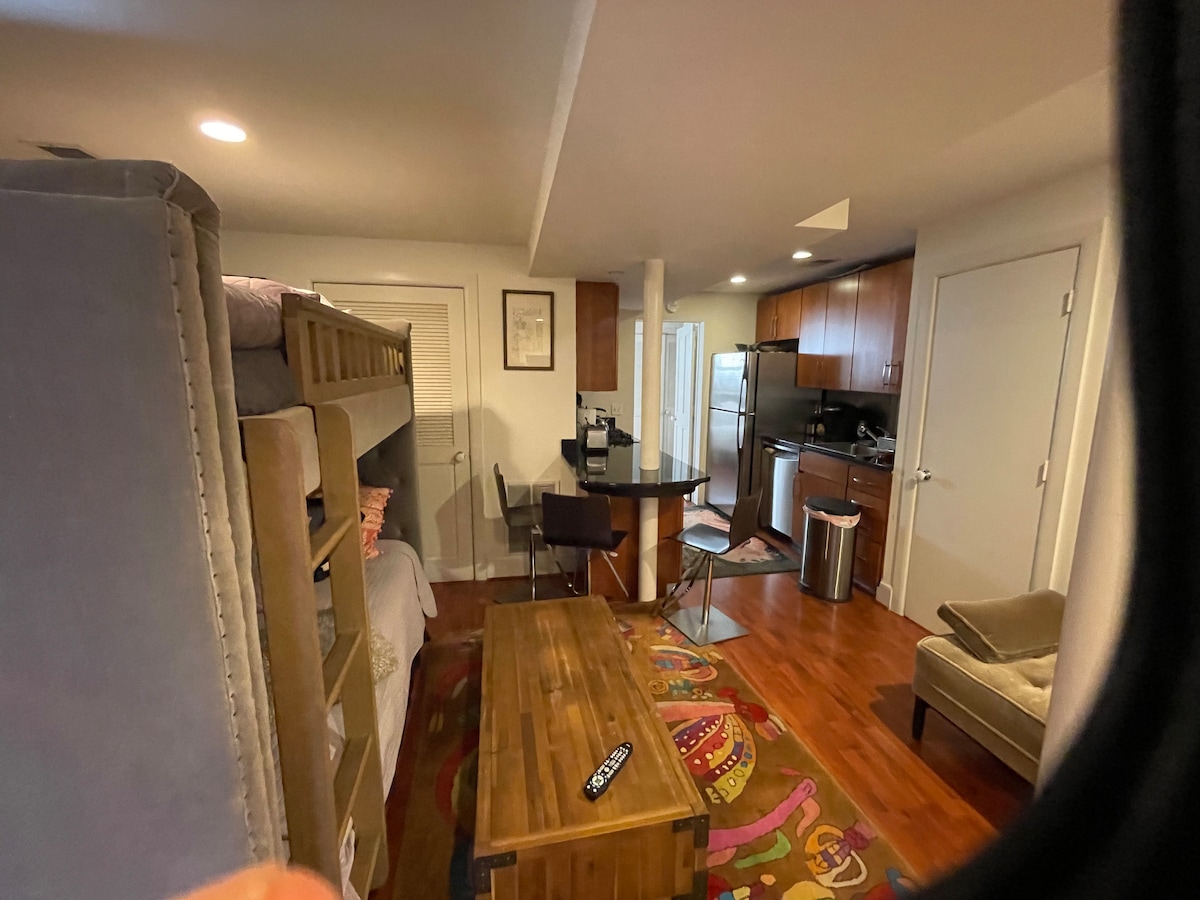 A compact living space includes a bunk bed, a wooden coffee table, and a small dining area. The kitchen features modern appliances, while warm wood tones and a colorful rug enhance the interior. Natural light fills the room through wall openings.