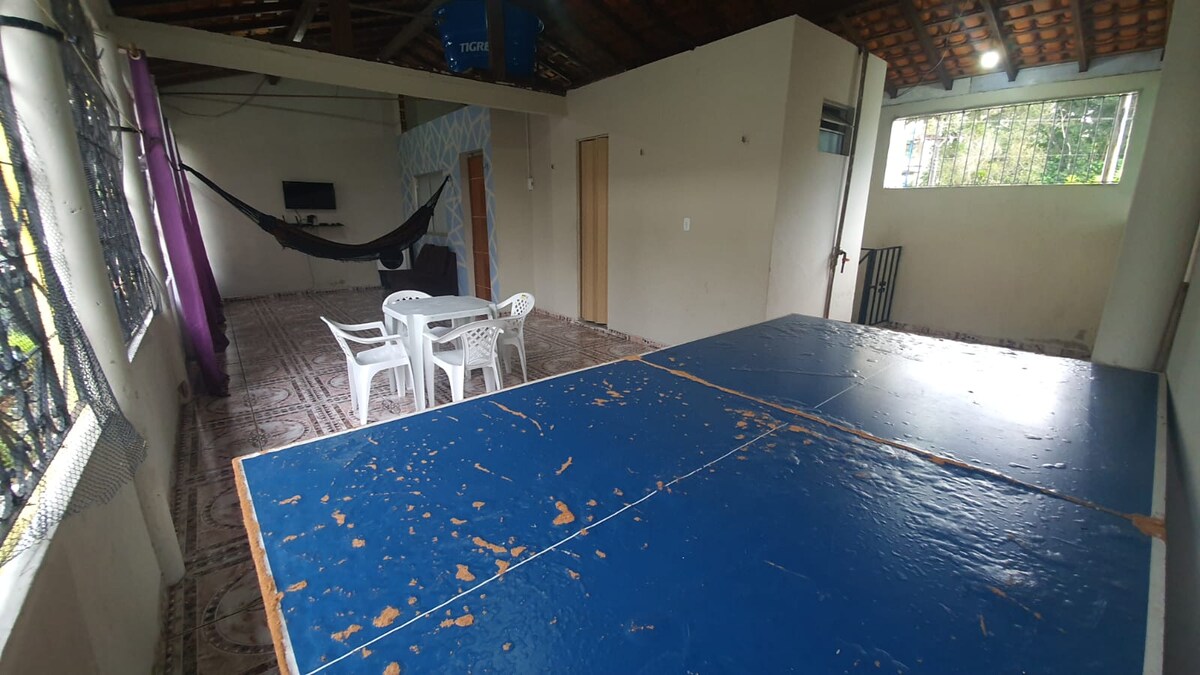 An open recreational space featuring a blue ping-pong table with visible wear marks. Nearby, four white plastic chairs surround a small table. A hammock is installed against the wall, and large windows allow natural light to fill the room.