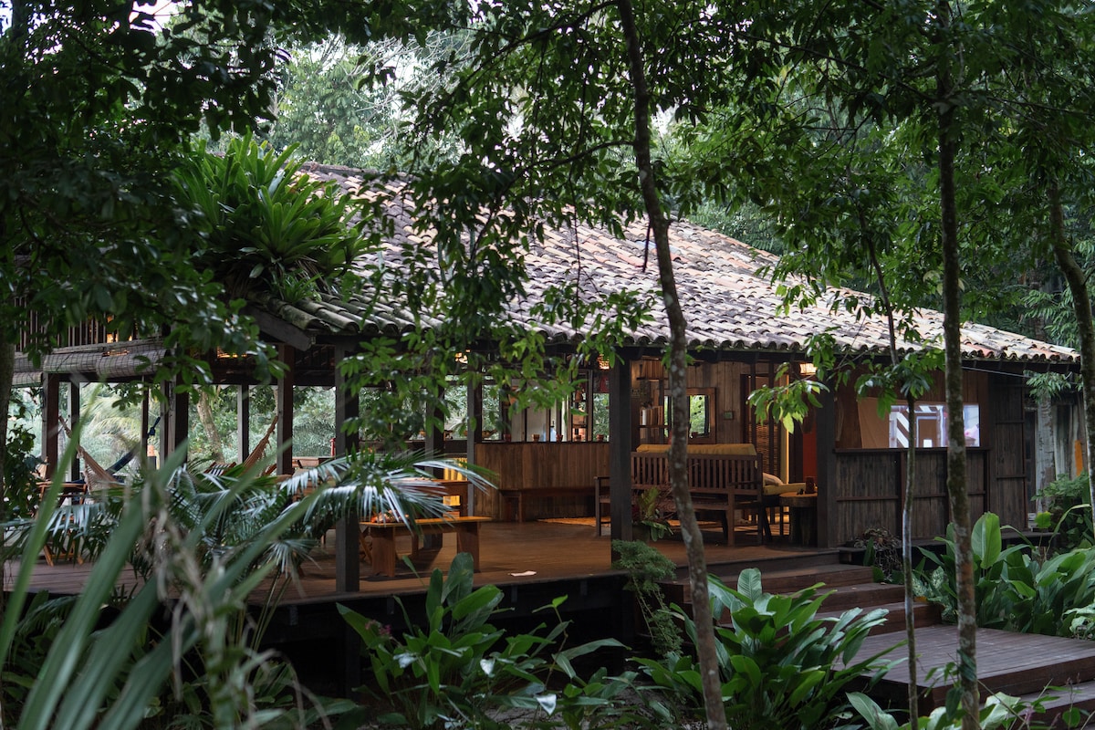 A spacious wooden bungalow is nestled among lush greenery in the tropical forest. The structure features a thatched roof and large windows, allowing natural light to illuminate the interior while blending harmoniously with the surrounding nature. Deck seating is visible on the outdoor patio.
