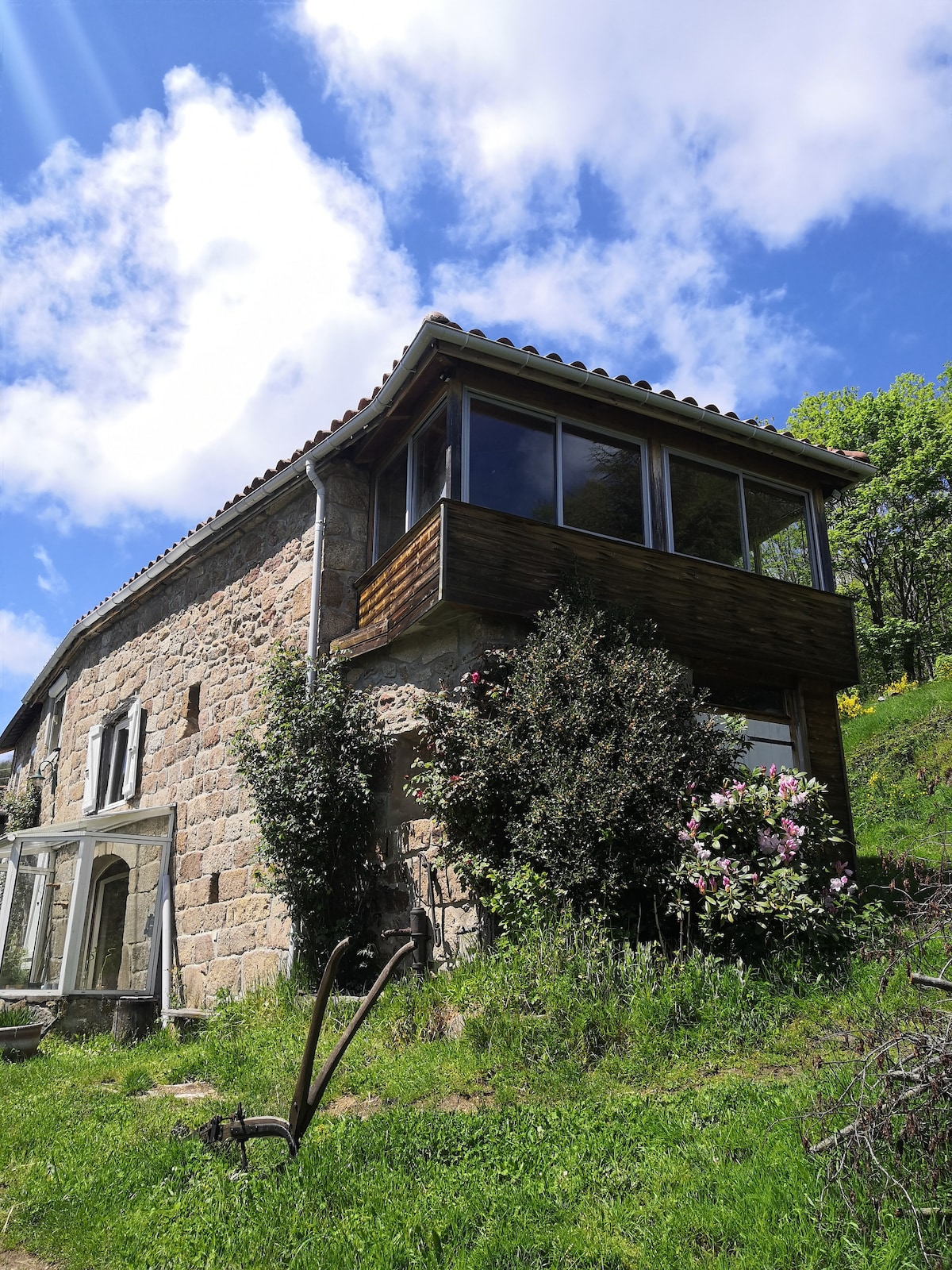 The exterior of a stone farmhouse is captured, showcasing lush greenery and flowering shrubs at its base. A spacious loggia with large glass windows is visible on the upper level, allowing for natural light. Blue skies and wispy clouds frame the structure.