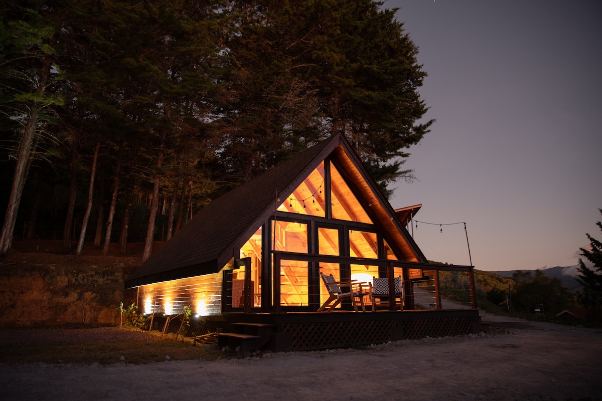 A modern chalet features wooden beams and large glass windows, showcasing a warm glow from interior lights. Surrounded by tall trees, the exterior includes a spacious deck with chairs, ideal for enjoying the serene forest ambiance. The mountain landscape is visible in the background.