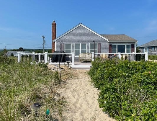 A charming cottage is set against a clear blue sky, with a white railing encircling a spacious deck. A sandy pathway leads to the entrance, flanked by lush greenery and beach grasses. The structure features a shingle exterior and a cozy chimney, enhancing its beachside appeal.