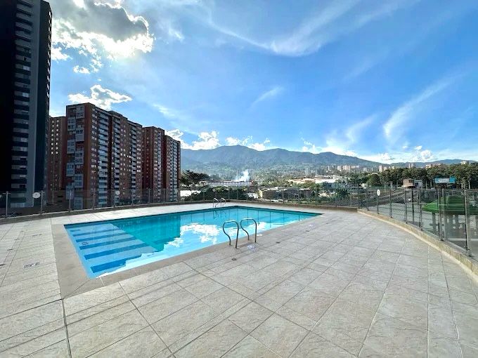 A clean and inviting swimming pool is surrounded by a spacious tiled area, offering stunning views of the nearby mountains and cityscape. The sky reflects vibrant blue hues, creating a serene atmosphere for relaxation and leisure.
