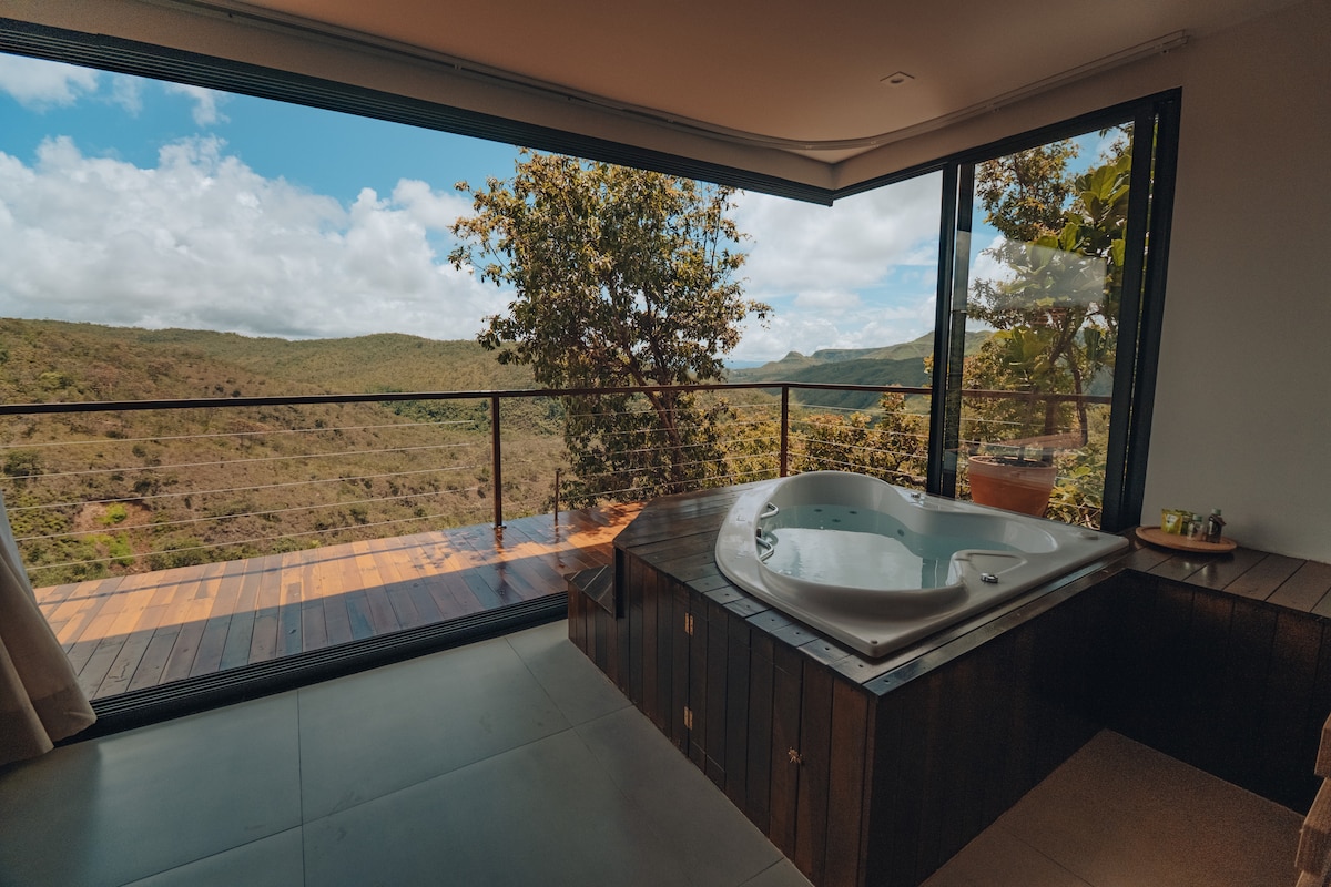 A spacious area featuring a hydromassage bathtub positioned next to large glass windows offering stunning views of the surrounding valleys. Natural light accentuates the modern design and wooden elements, creating a serene atmosphere for relaxation.