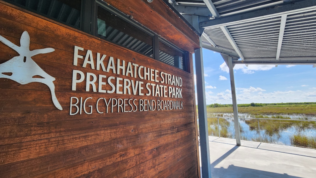 A wooden sign reading 'Fakahatchee Strand Preserve State Park' is prominently displayed, indicating the entrance to the boardwalk. The background features a wide, open vista of wetland grasses under a blue sky with wispy clouds, emphasizing the natural beauty of the area.