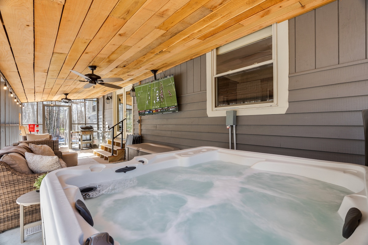 A spacious hot tub is positioned on a covered porch, with a wooden ceiling overhead. Nearby, a wall-mounted TV displays programming, and comfortable seating is arranged for relaxation. Natural light filters through nearby windows, creating a serene atmosphere.
