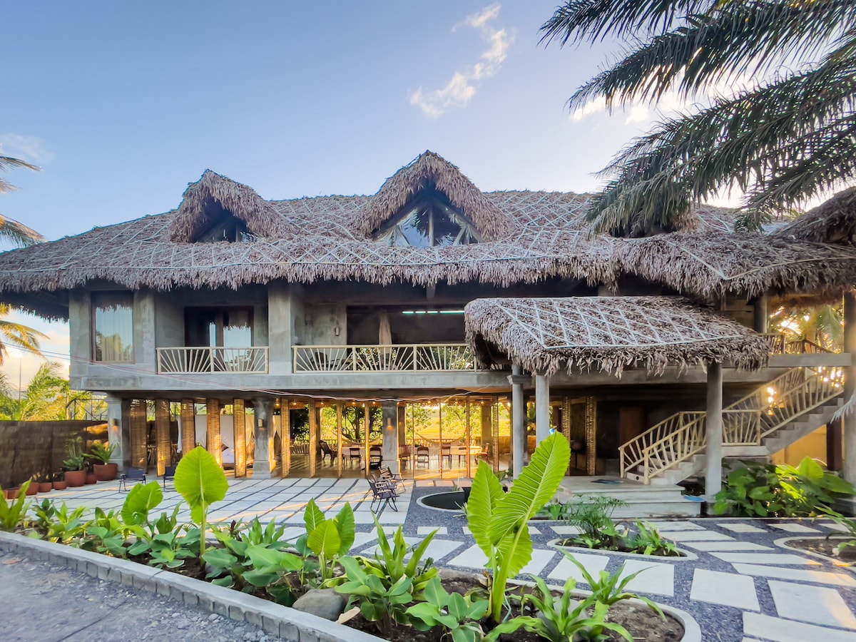 A Balinese-inspired villa features a thatched roof and a harmonious blend of natural materials. The expansive structure is surrounded by lush greenery, with large windows allowing for ample natural light. A manicured pathway leads to a beautifully landscaped garden area.