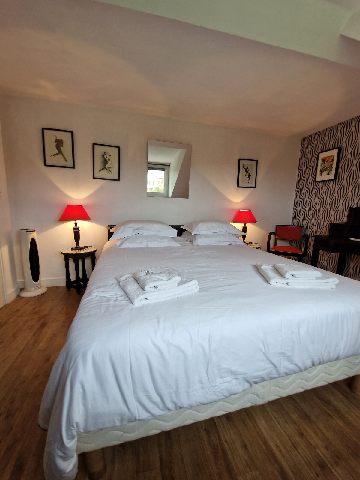 A welcoming bedroom features a spacious bed with crisp white linens and two neatly folded towels placed at the foot. Light is provided by bedside lamps on black tables, complemented by artwork on the walls. A desk and chair are visible in the corner.