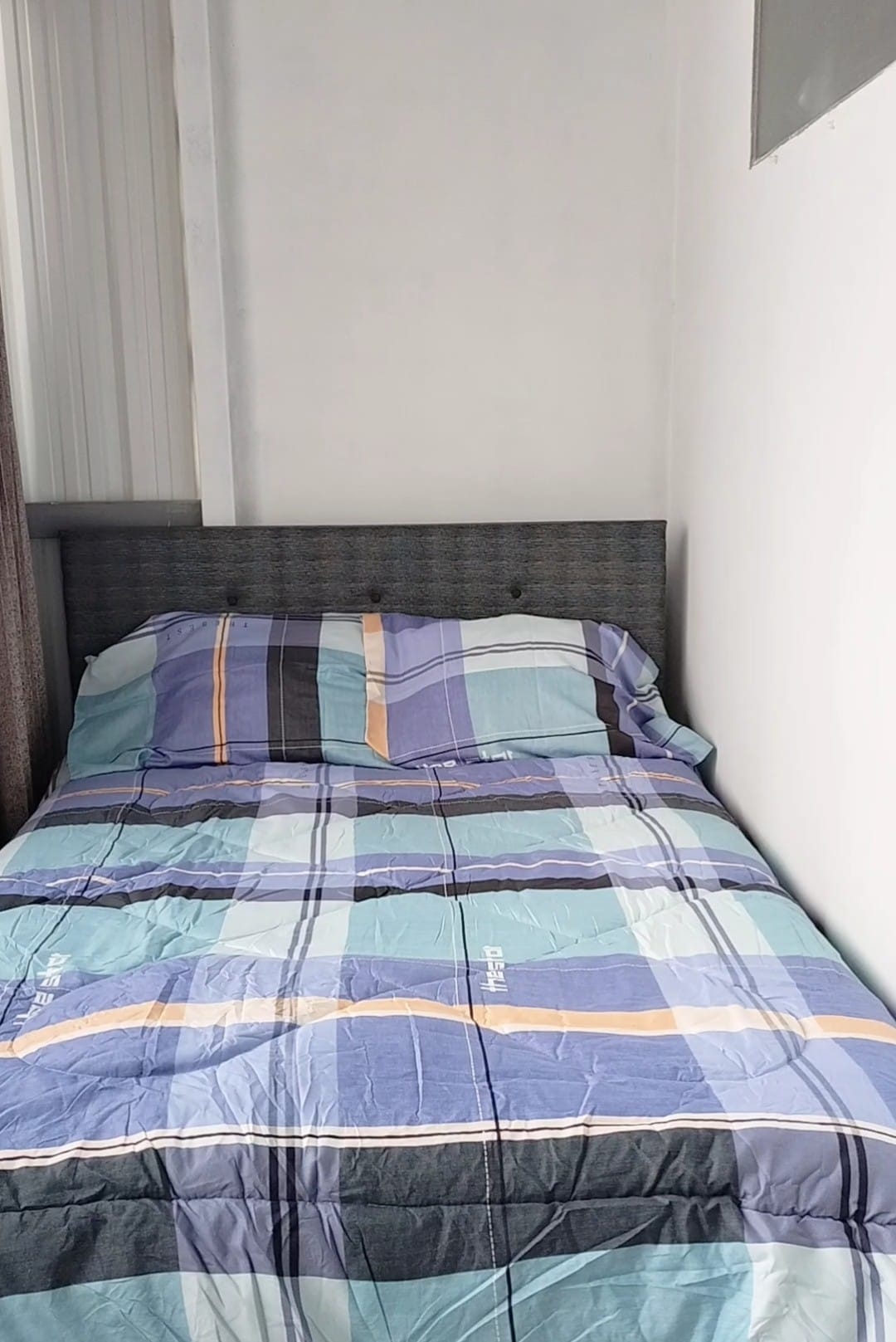 A compact sleeping area features a neatly made bed with a patterned blanket in shades of blue and green. The headboard is upholstered, and the wall beside the bed is adorned with a light color, creating a clean and simple look.