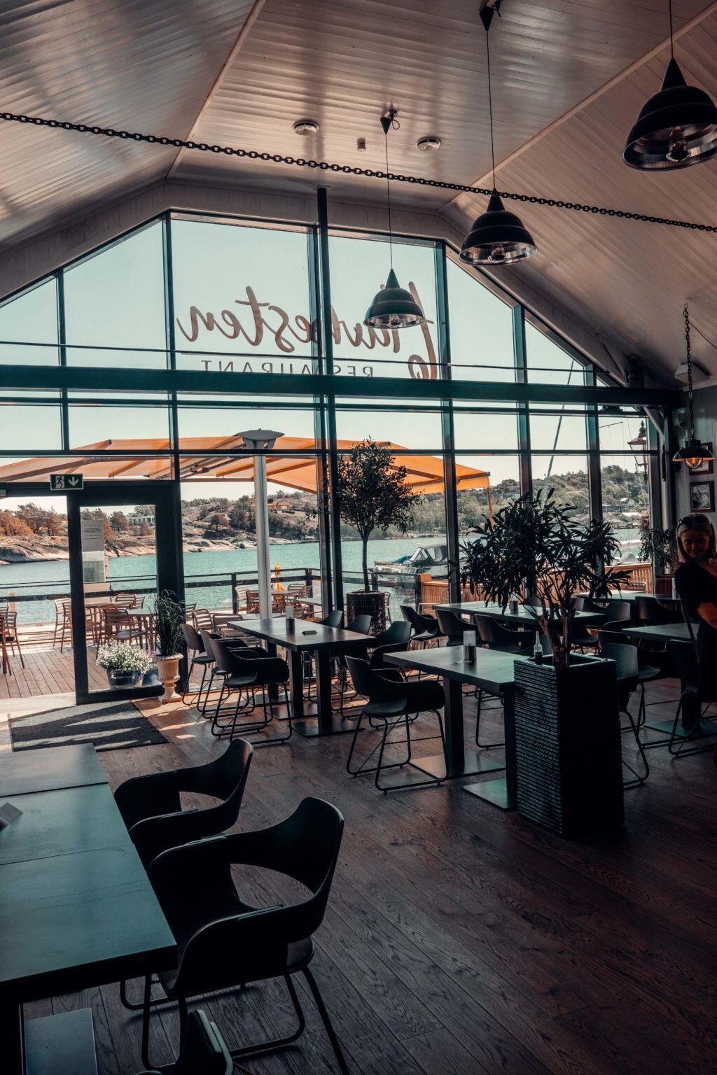 A modern dining space features large windows offering views of the water. Indoor plants and stylish lighting fixtures enhance the atmosphere. Tables and chairs are arranged to allow for comfortable dining, with an outdoor terrace visible through the glass, providing access to natural light.