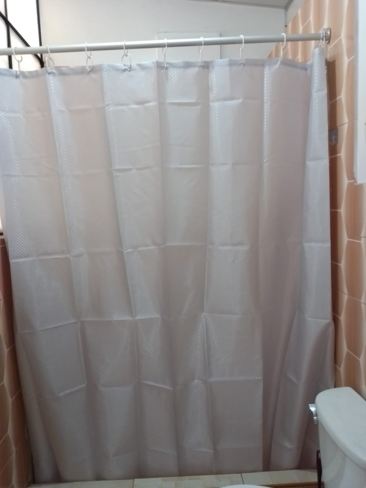 A straightforward bathroom setting is presented, showcasing a shower area with a light-colored shower curtain. The curtain features a simple design and hangs from a metal rod. A white toilet is positioned adjacent to the shower space, complementing the tiled walls.