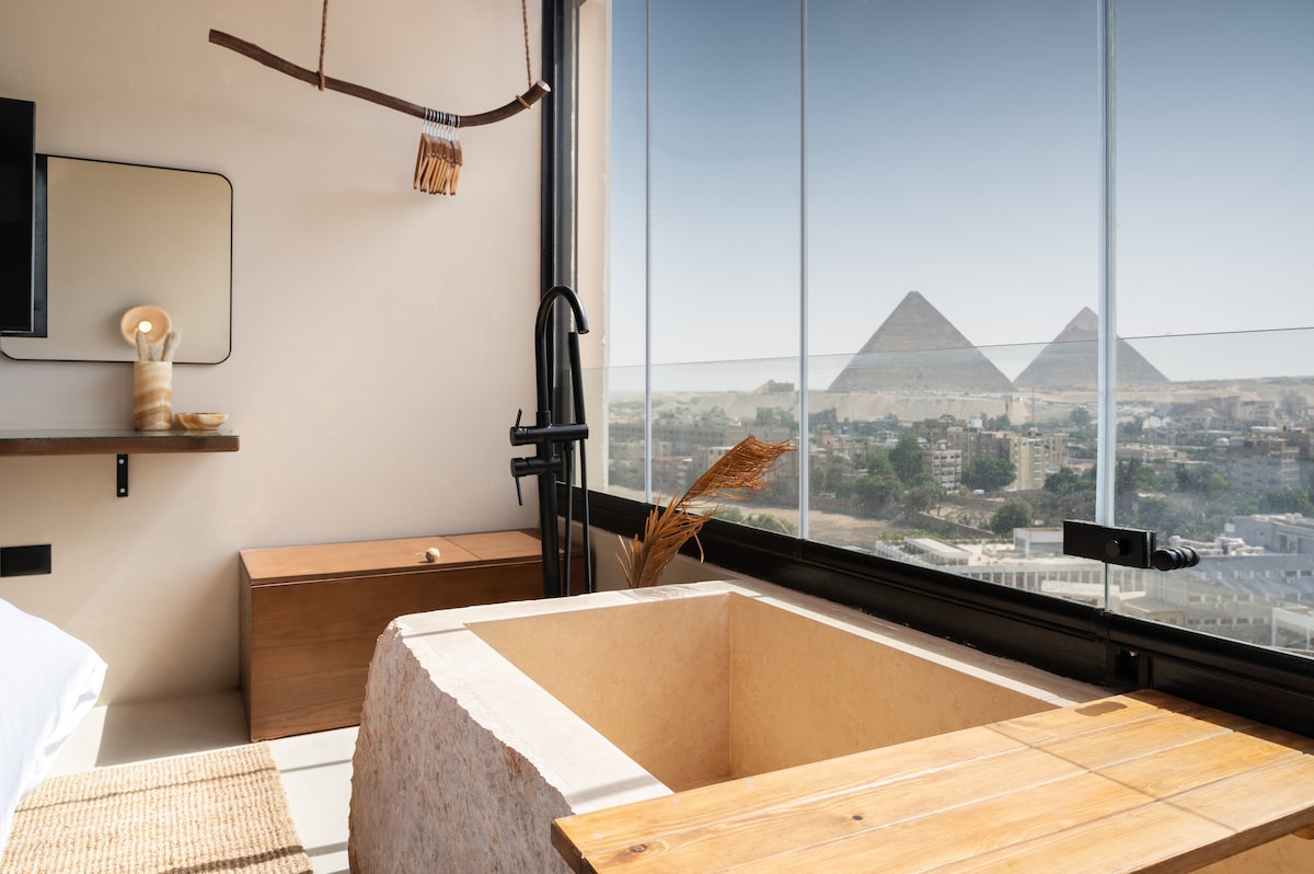 A full-sized bathtub, crafted from natural materials, is positioned beside expansive windows offering unobstructed views of the Giza Pyramids. Minimalistic design elements, including a sleek faucet and wooden accent, complement the serene atmosphere of the room.