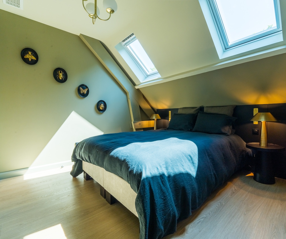 A sleeping area with a spacious bed dressed in dark bedding is positioned under sloped ceilings. Two skylights bring natural light into the room. Three decorative bee wall hangings add a touch of character, while a small nightstand is located beside the bed.