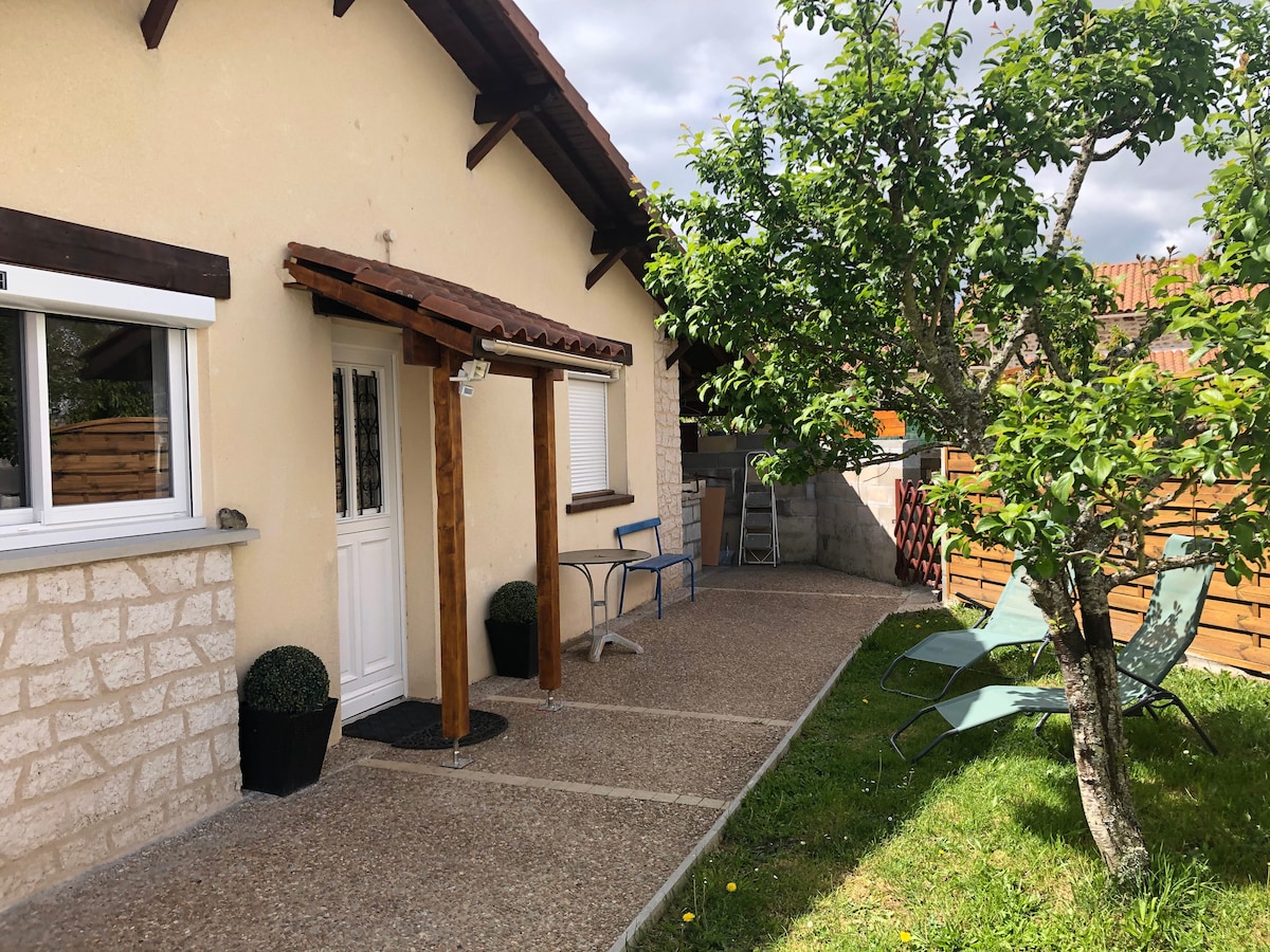 The exterior of the home is shown, featuring a welcoming entrance with a wooden overhang and a white door. Two lounge chairs are placed on the grassy area, alongside a small tree and decorative potted plants, creating a serene outdoor space.