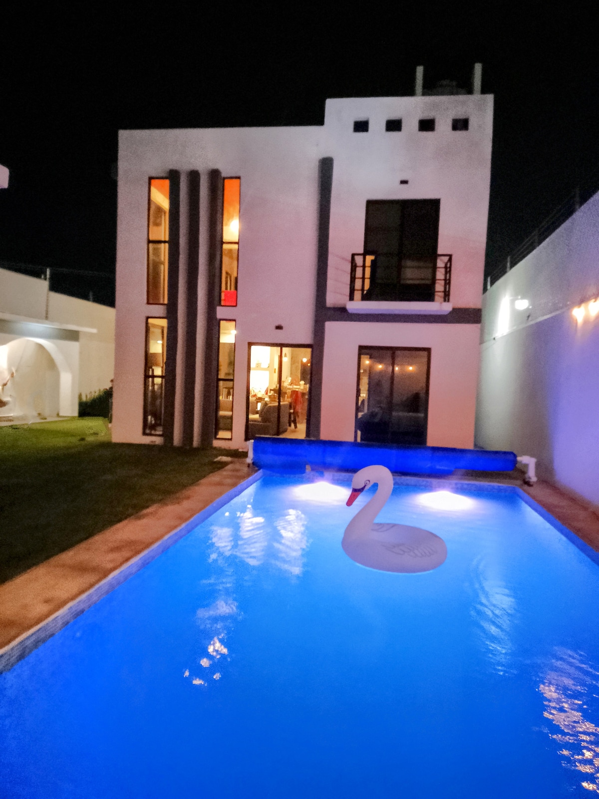 The exterior of a modern two-story house is presented at night, featuring illuminated glass doors that reveal warm interior lighting. A sparkling pool occupies the foreground, with a swan-shaped float gently resting on the water's surface, surrounded by soft blue lighting.