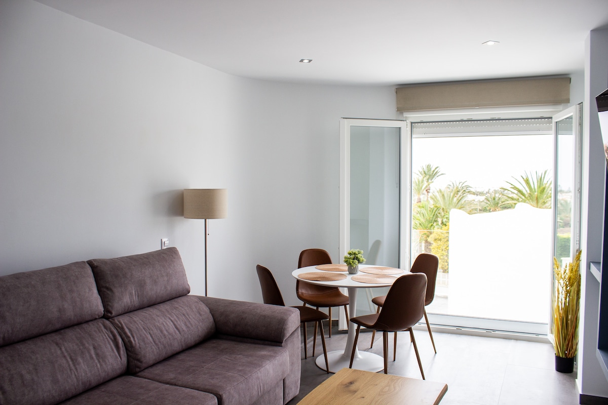 A spacious living area features a contemporary gray sofa, complemented by a wooden coffee table. A dining table with four brown chairs is positioned near a large window that opens to a view of greenery. Natural light enhances the fresh, bright ambiance.