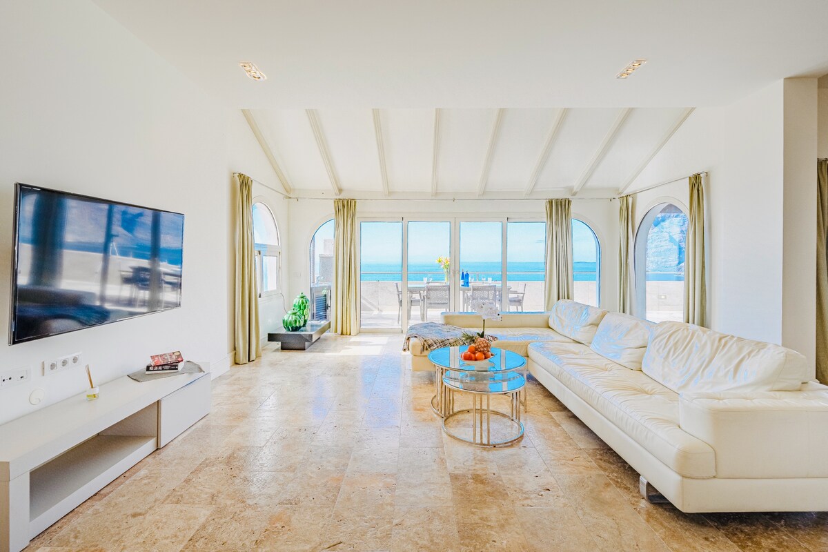 A spacious living area is filled with natural light, featuring large arched windows that offer a view of the ocean. A sectional sofa is positioned around a coffee table, complemented by decorative plants. A wall-mounted television is visible, enhancing the modern ambiance.