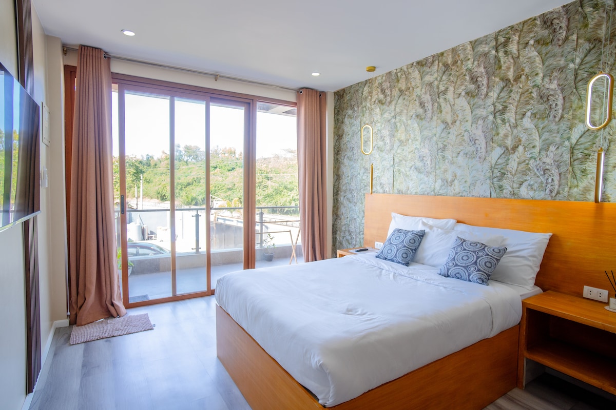 A bright room features a spacious bed with white linens and decorative pillows. Wood accents complement the modern decor. Large glass doors open to a private balcony, allowing natural sunlight to fill the space and offering views of the lush surroundings.
