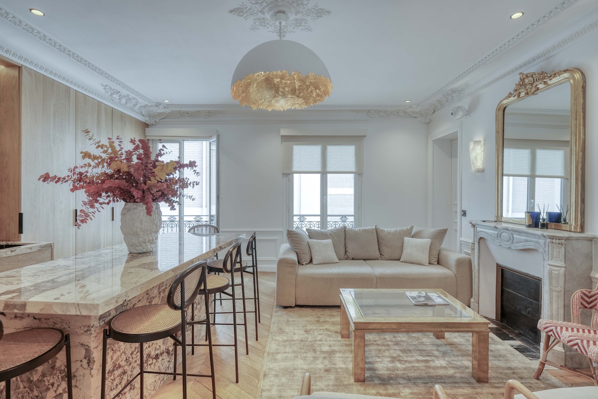 An elegantly appointed living space features a spacious beige sofa and a marble coffee table. A large floral arrangement sits in a vase on the marble island, complemented by sleek bar stools. Natural light floods the room through the large windows, highlighting intricate ceiling details.