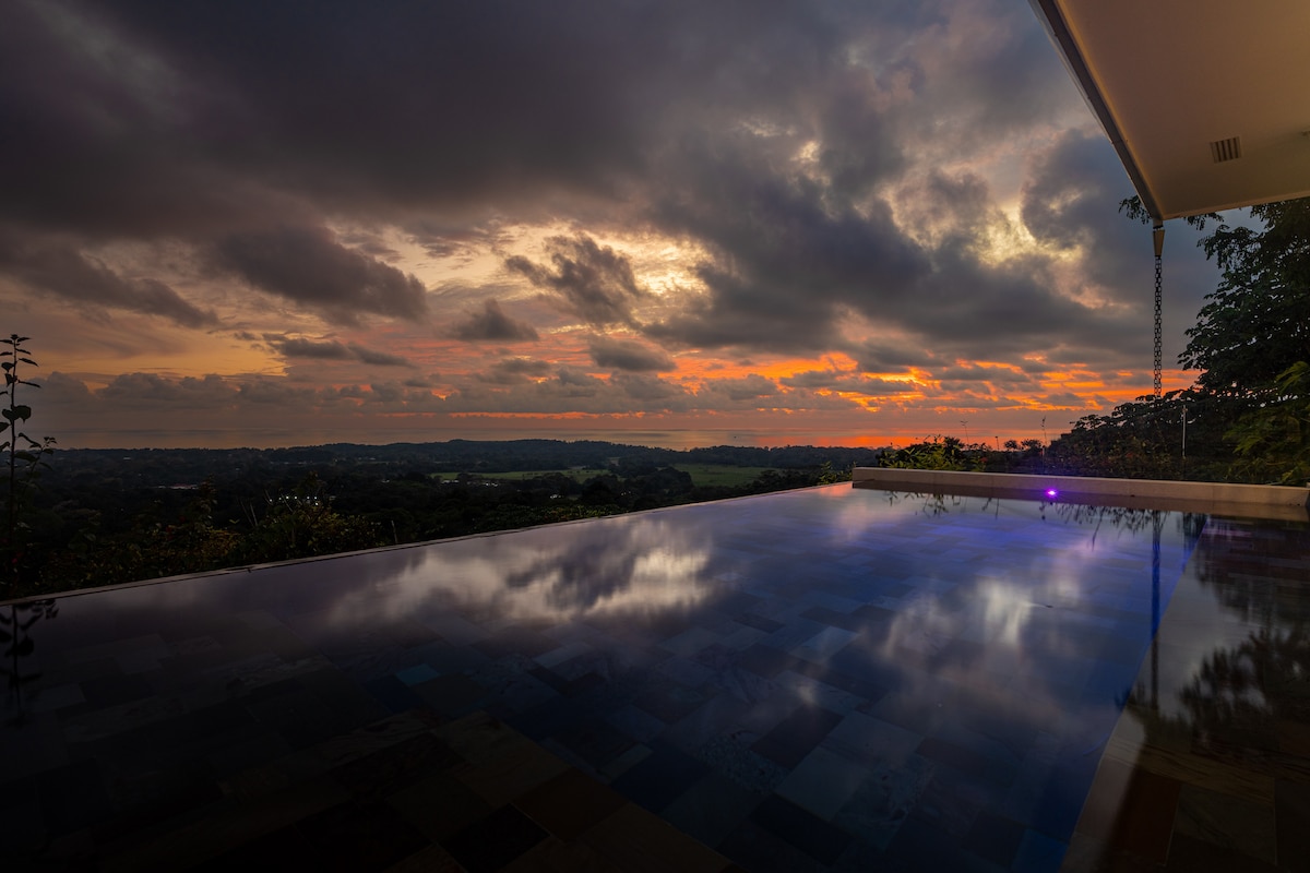 A tranquil infinity pool reflects the colorful hues of a sunset over Uvita. The landscape showcases lush greenery and distant hills, framed by a serene sky filled with clouds. Soft lights near the pool add to the peaceful ambiance.