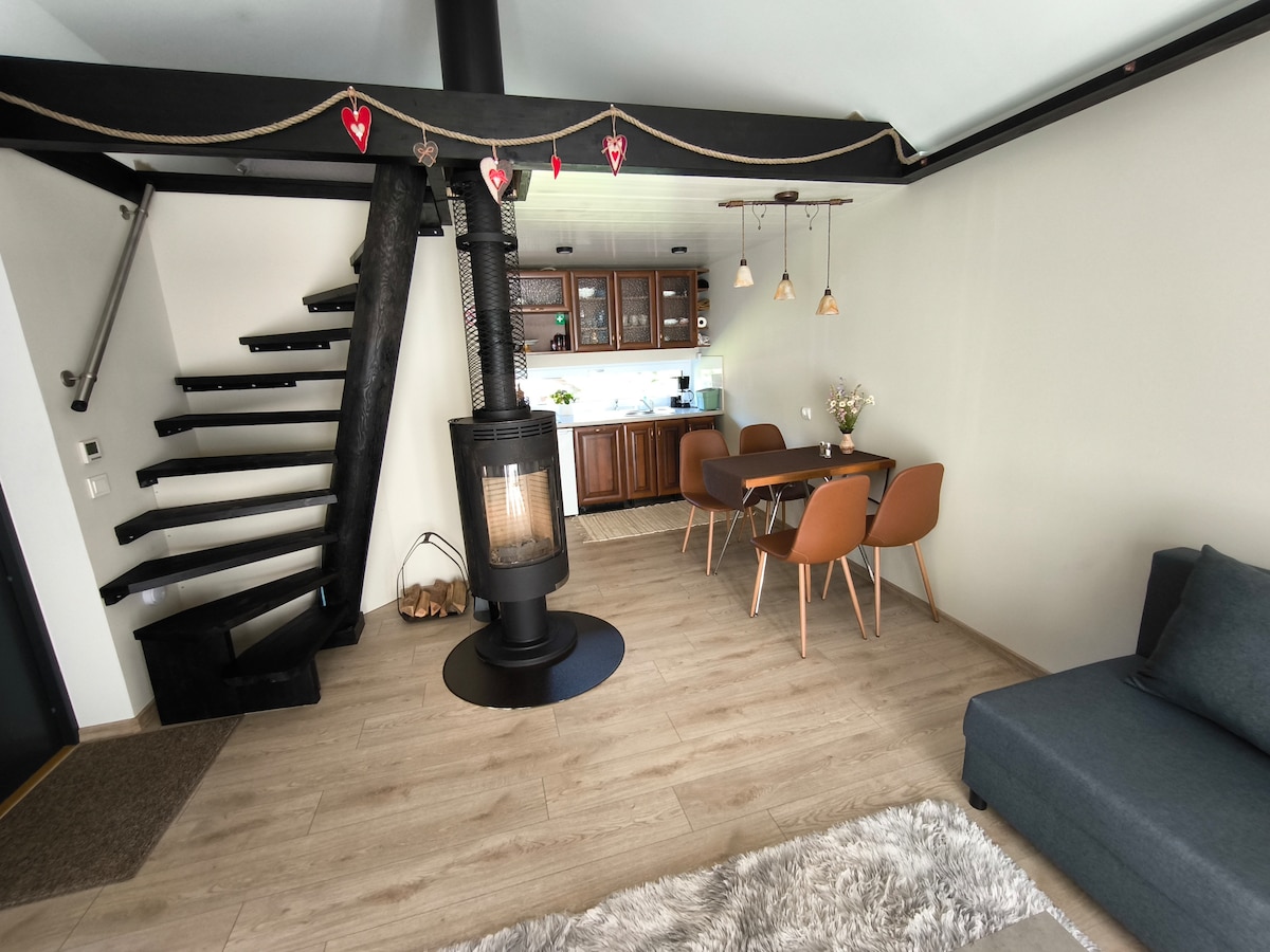 The living area features a stylish dining space with a wooden table and four chairs, illuminated by pendant lights. A modern stove provides warmth, while a comfortable sofa and a staircase lead to the upper level. Light-colored flooring enhances the welcoming ambiance.