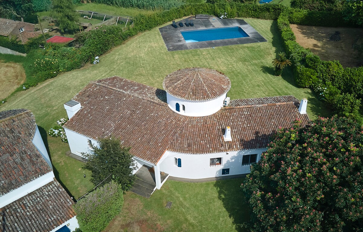 An aerial view captures the charming white house with a circular roof, surrounded by lush green gardens. The landscape includes a private swimming pool and various outdoor spaces, all set in a tranquil environment.
