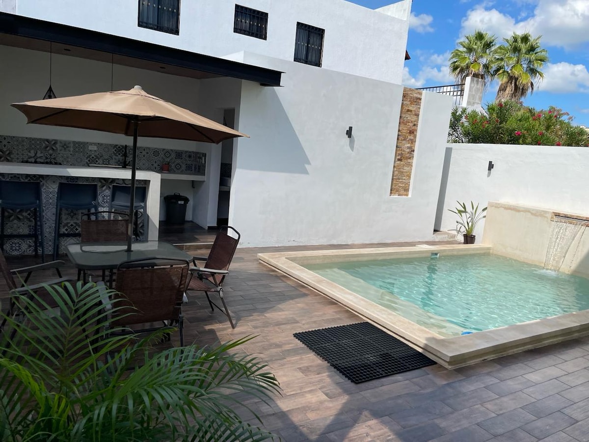 A shaded patio area features a table with four chairs and an umbrella, providing a comfortable outdoor dining space. A small pool is visible, surrounded by tiles, with water gently flowing over a short wall. Lush greenery and palm trees frame the space.