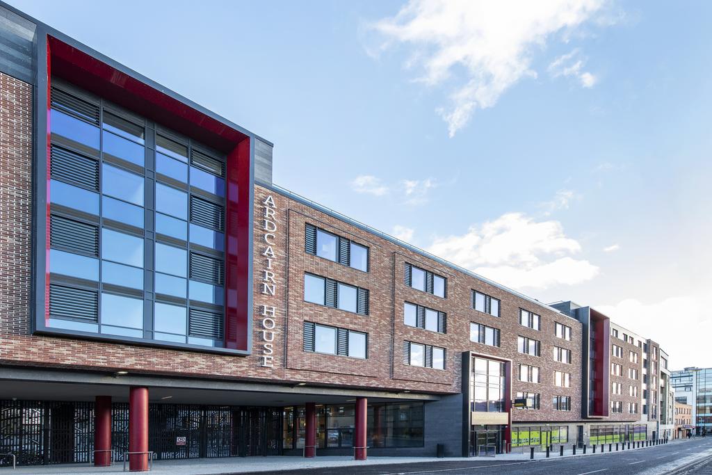 A modern student residence building is depicted, featuring a mix of red and brown brick exteriors. Large glass windows create a bright, inviting facade. The entrance is marked by the name 'Aldcairn House,' prominently displayed, while the surrounding area shows a well-maintained pathway.
