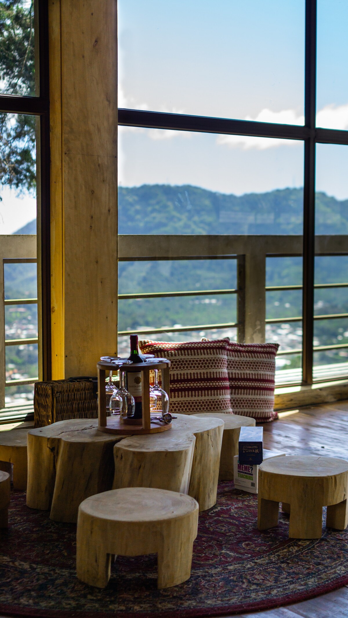 A wooden table constructed from natural logs is set in a cozy corner, accompanied by smaller log stools. A view of the mountains is visible through large windows, while a woven basket and decorative pillows add comfort to the space.
