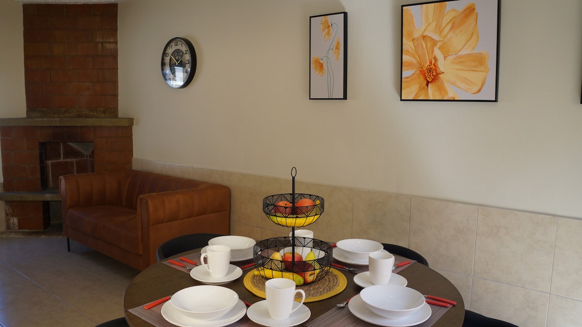A dining area is shown with a table set for four, featuring dishes, cups, and utensils. A fruit basket sits at the center. The walls are adorned with floral artwork, while a comfortable brown sofa is positioned nearby, adding to the inviting environment.