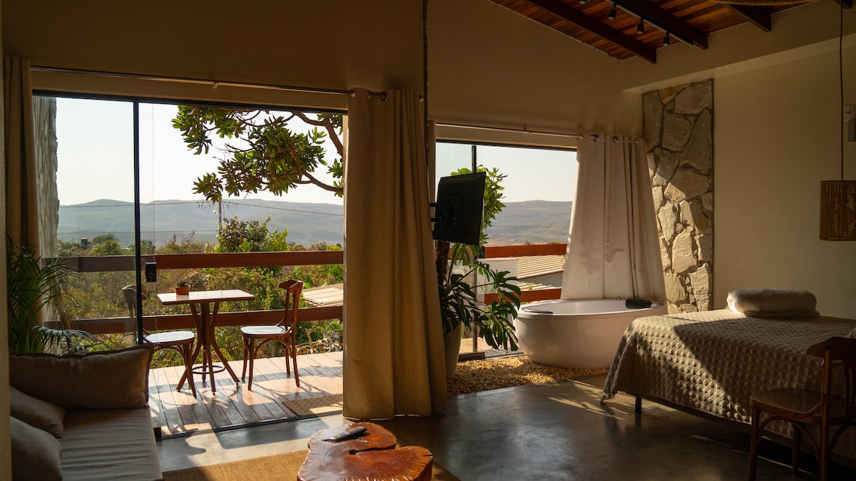 A bright interior features large glass doors opening to a scenic view of the landscape. A spacious bed and a cozy seating area are complemented by natural light. A small table with chairs sits on the private terrace, enhancing the connection to nature.