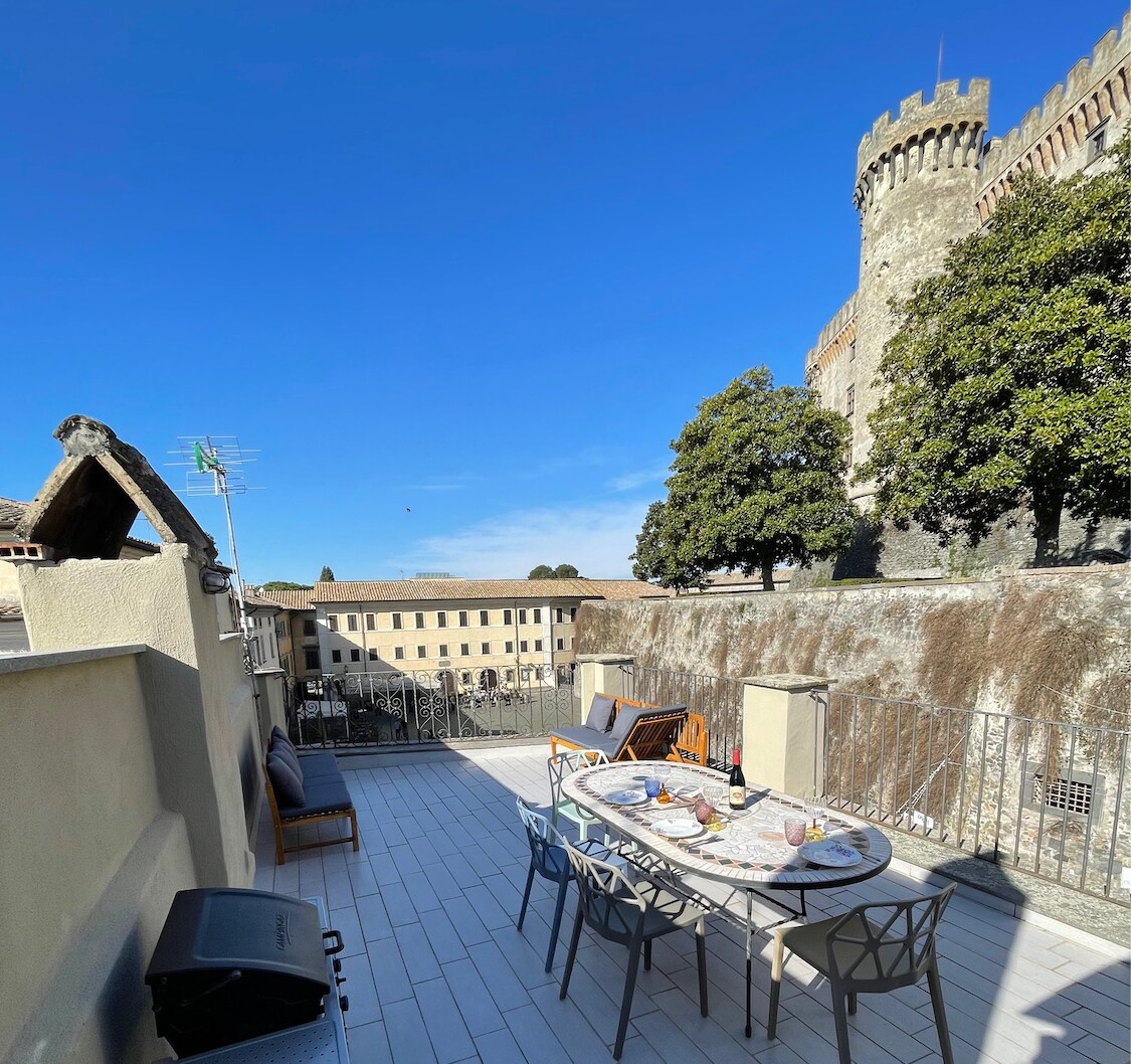 A private terrace is showcased with a dining table and chairs, overlooking the historic castle and surrounding buildings. The clear blue sky enhances the open feel, while greenery provides a natural touch. A grill is also visible, offering potential for outdoor cooking.