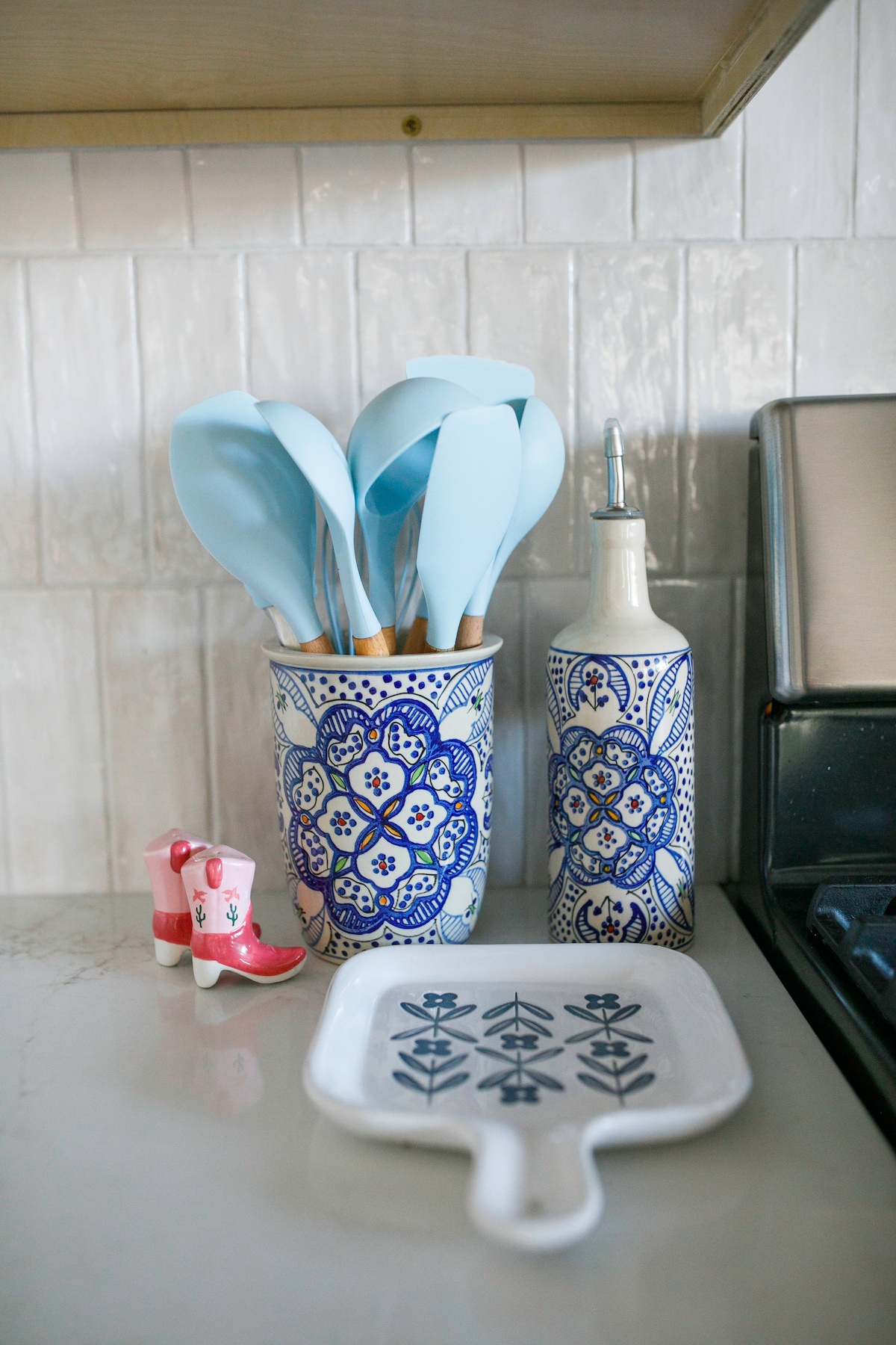 A collection of kitchen utensils is displayed in a decorative ceramic holder, featuring blue floral patterns. The utensils are blue silicone spatulas with wooden handles. A matching bottle for oil or vinegar and a small ceramic dish with a geometric design are also present, alongside a whimsical pair of pink and white toy boots.
