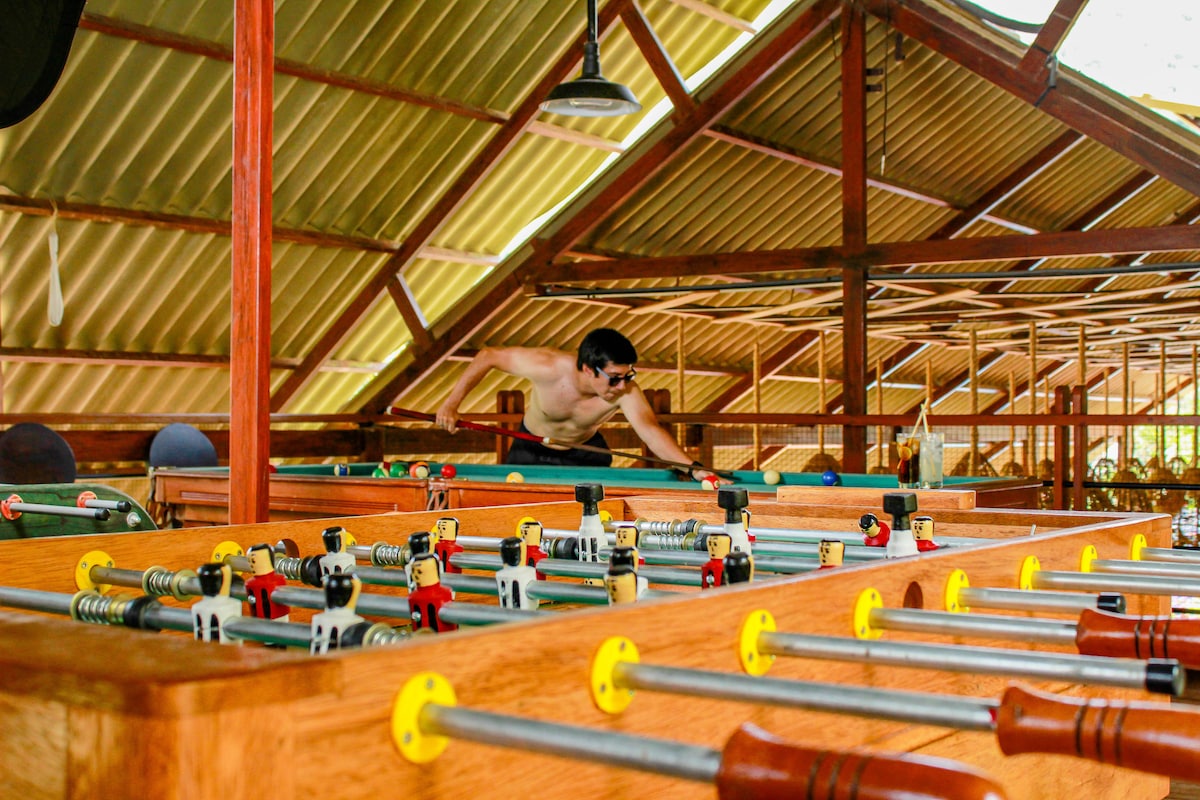A spacious game room is featured, showcasing several foosball tables crafted from wood. A player is seen engaging in the game, with the room's high ceiling and ample natural light enhancing the atmosphere.