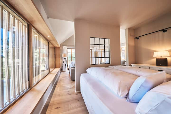 Über die modernen Holzlamellen blickt man beim Aufwachen bis zu den Bergen. Wie in einer Hotelsuite ist dieses Zimmer geöffnet zum Wohnraum. Gleich neben dem Bett befindet sich der Eingang zum eigenen Natursteinbad mit Regendusche.