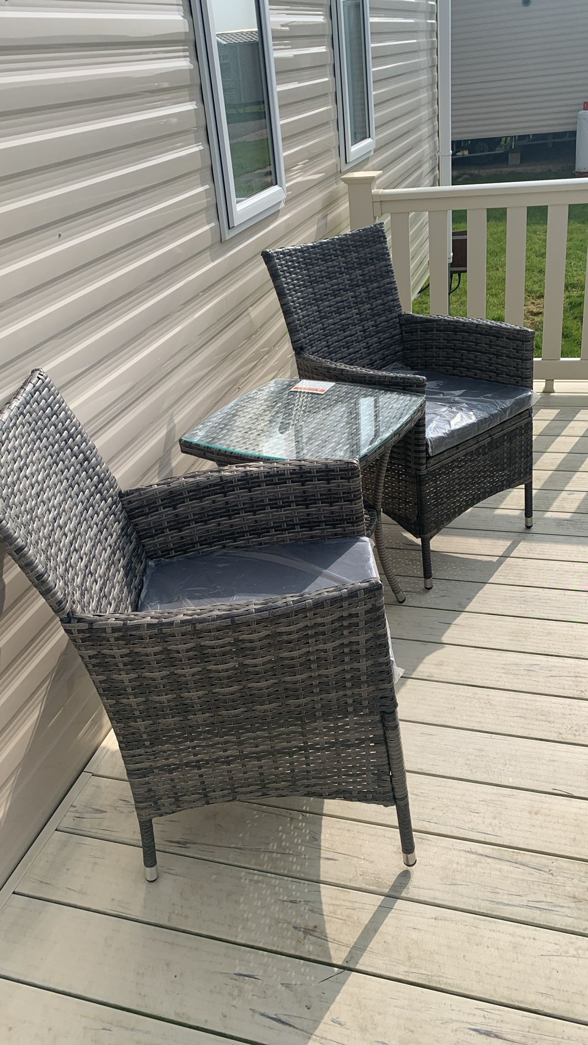 A small outdoor seating area is present, featuring two woven chairs with soft cushions positioned around a glass-top table. The space is surrounded by a wooden railing, and natural light illuminates the setting from nearby windows.