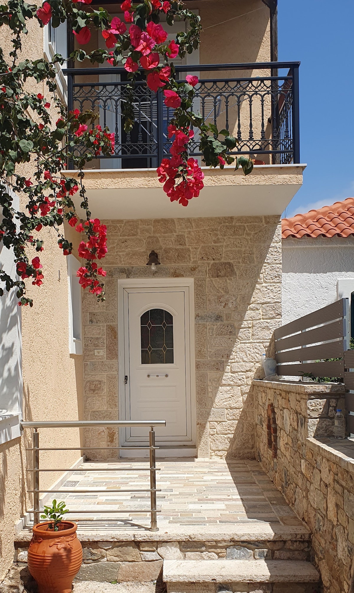 A charming entrance features a stone pathway leading to a white door, framed by a decorative arch. Colorful bougainvillea vines cascade down from the balcony above, contrasting with the sunlit beige walls of the building. A small potted plant adds a touch of greenery to the space.