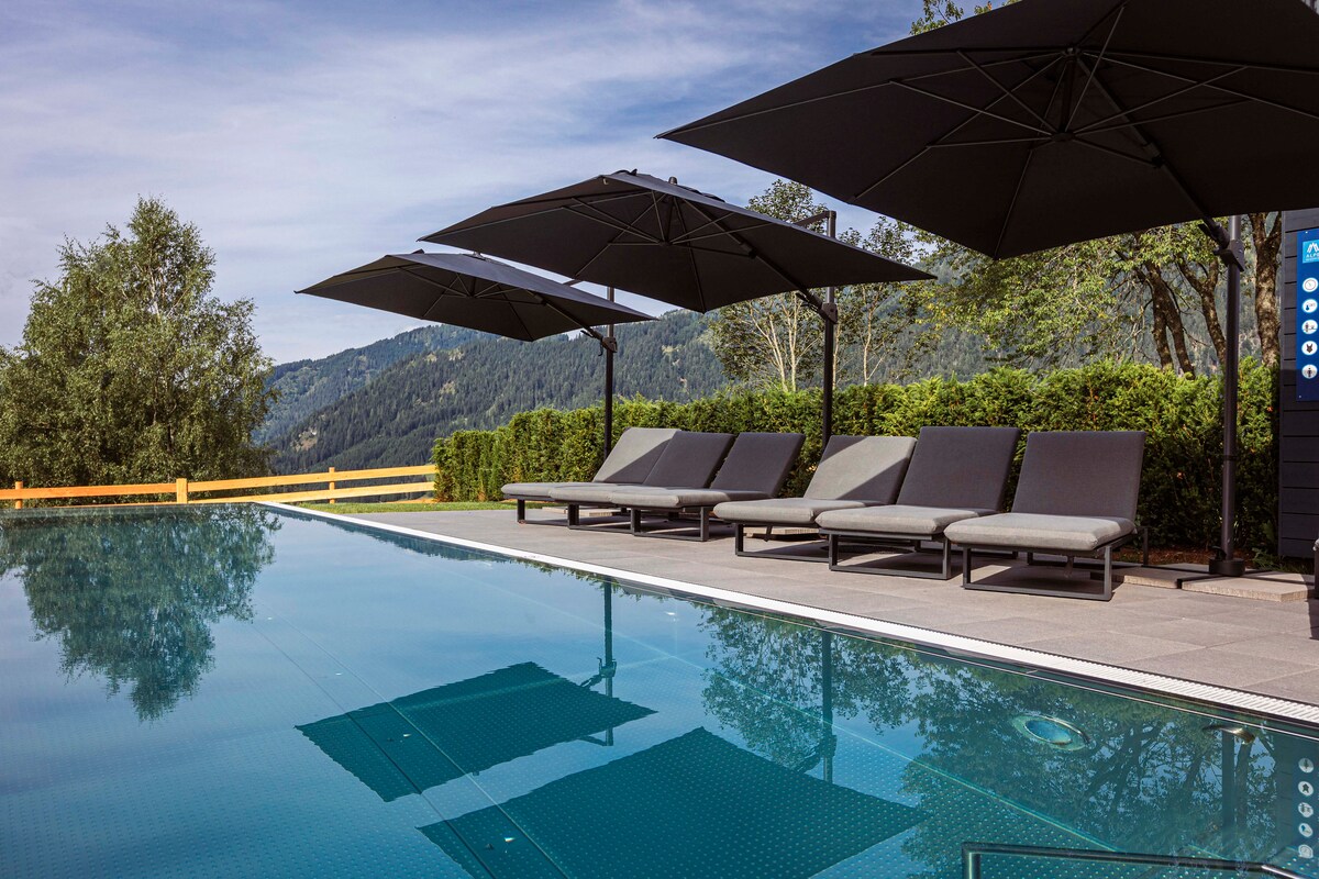A heated outdoor infinity pool is surrounded by sleek lounge chairs positioned under large umbrellas. Lush greenery and a wooden fence border the area, with mountain views visible in the background, enhancing the serene atmosphere of the setting.