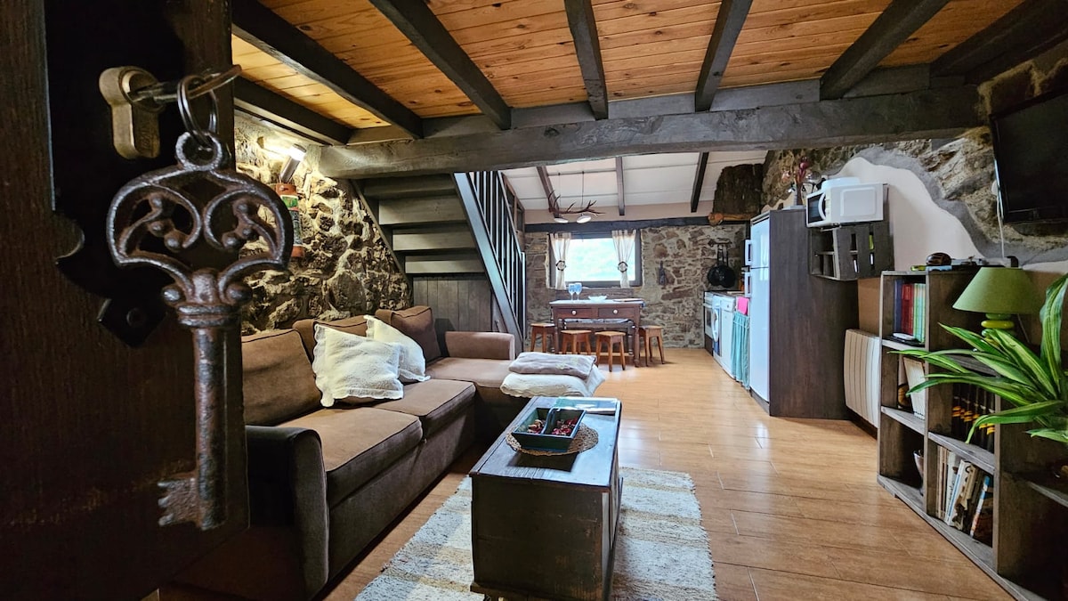 A cozy interior showcases a combination of stone walls and wooden beams. A comfortable sofa is positioned in front of a wooden coffee table, while a dining area is visible in the background. Natural light streams in through a window, creating a warm ambiance.
