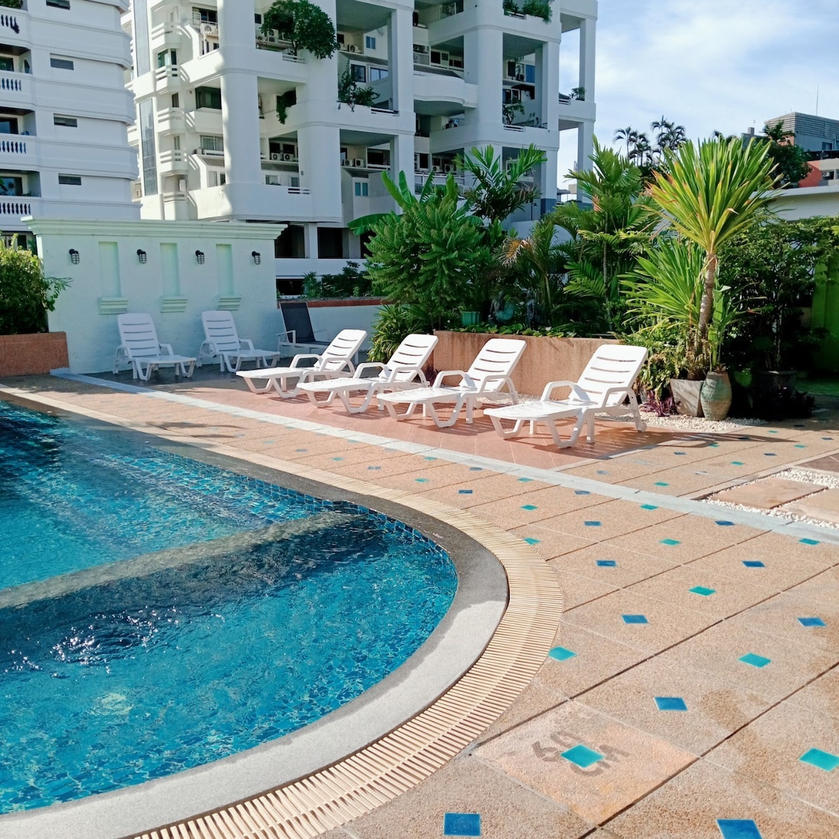 A swimming pool area is depicted, featuring a curved edge with blue water. Several white lounge chairs are positioned around the pool, surrounded by green plants and vibrant landscaping. A multi-story building is visible in the background under a clear sky.