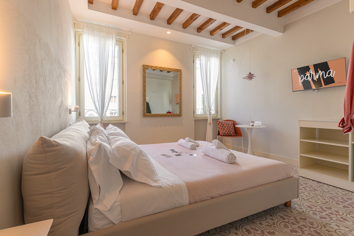 A bright and airy sleeping area is furnished with a spacious bed, featuring neatly arranged white linens and towels. Natural light filters through two windows, complementing the room's soft color palette. A wall-mounted smart TV and a stylish mirror enhance the modern design.