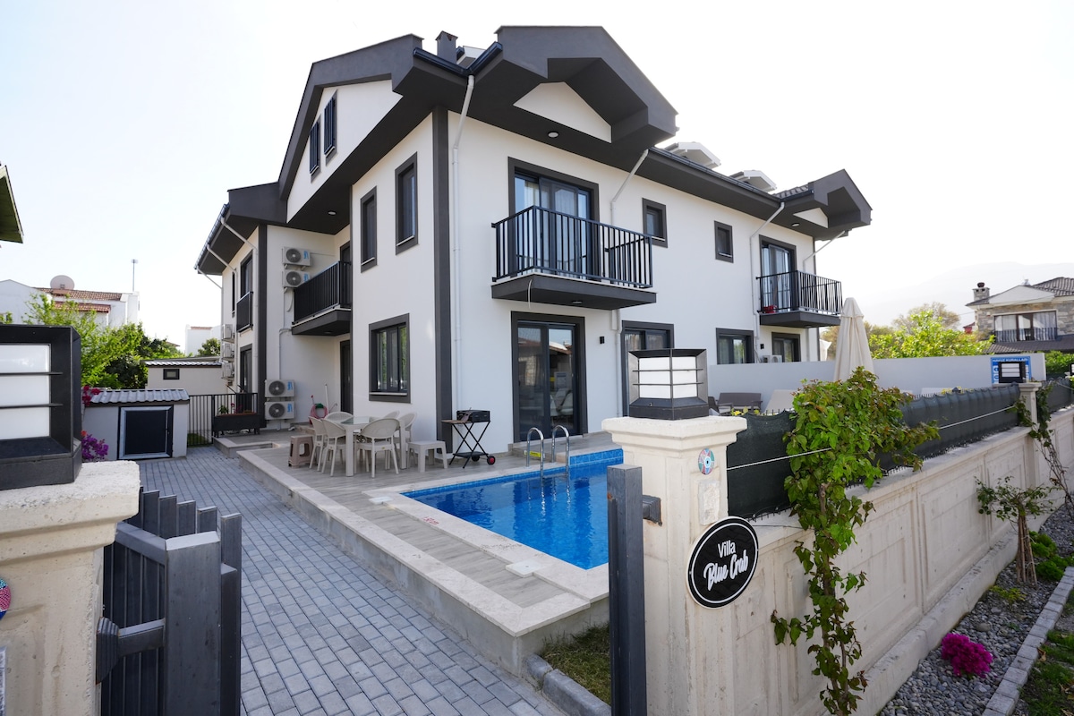 A modern villa exterior is presented, featuring a private pool and surrounding terrace. The structure has multiple balconies and large windows, allowing natural light to flood the interior. A neatly paved pathway leads from the entrance, framed by greenery and outdoor seating.