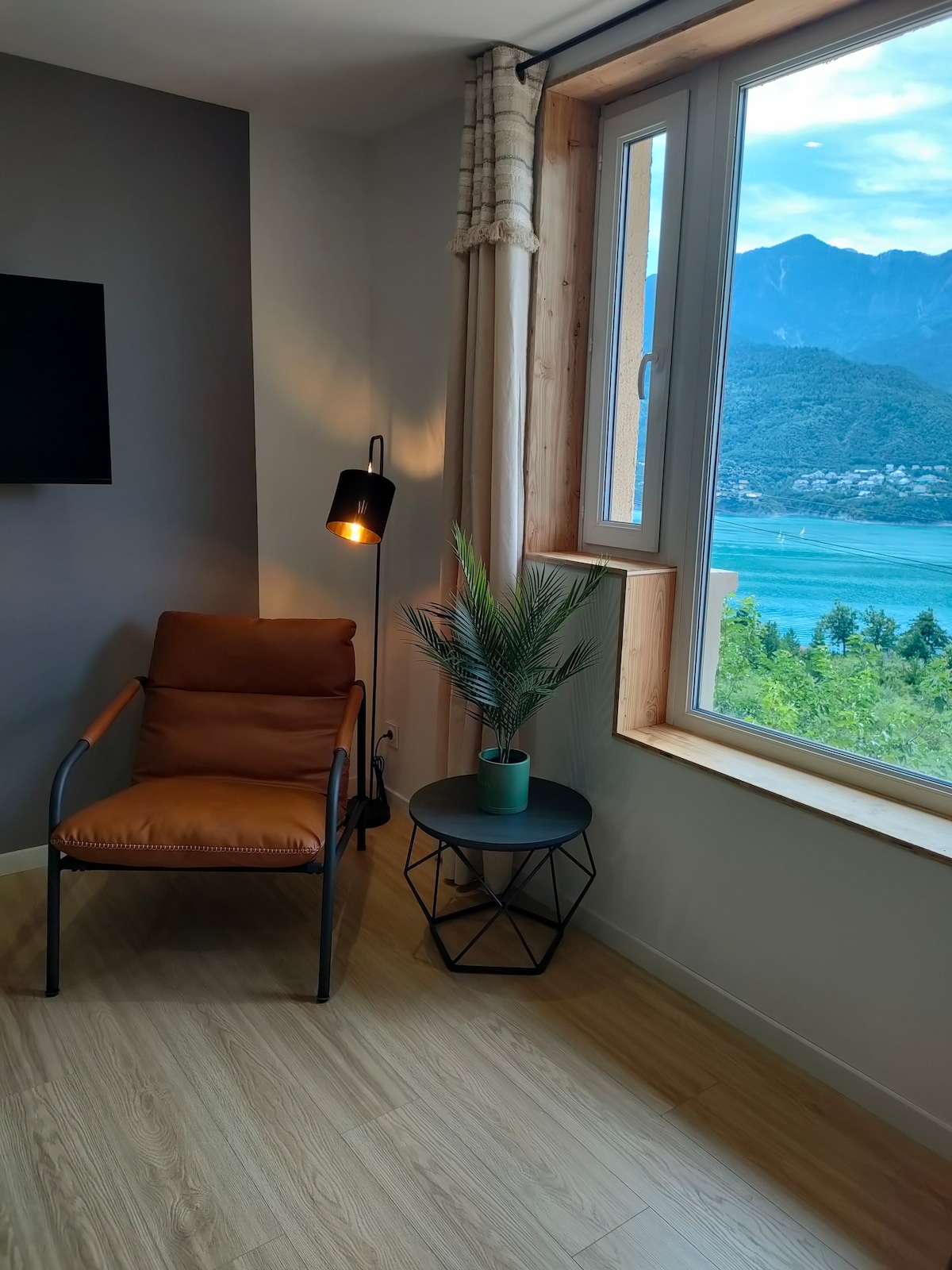 A corner of the room features a comfortable chair with a warm light beside it, positioned next to a small side table. A potted plant adds a touch of greenery, while large windows allow for a clear view of the lake and mountains outside.