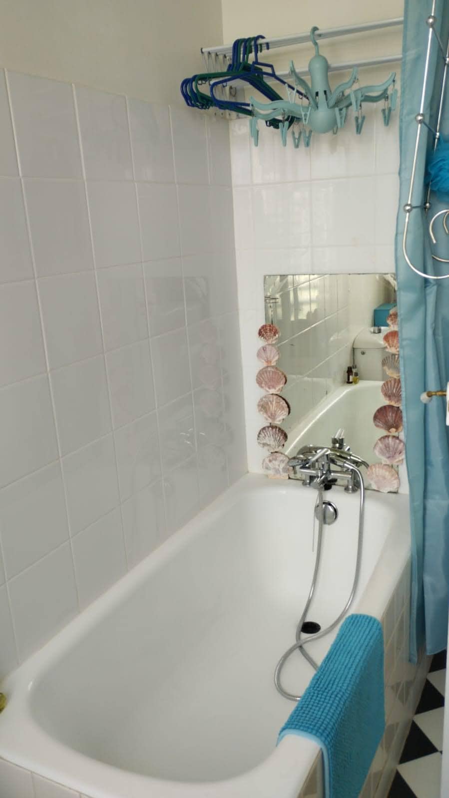 A bright bathroom containing a white bathtub with a handheld shower attachment is visible. A mirror framed with shells reflects the space, while blue accents, including a shower curtain and towel, add color. The tiled floor features a black and white checkerboard pattern.